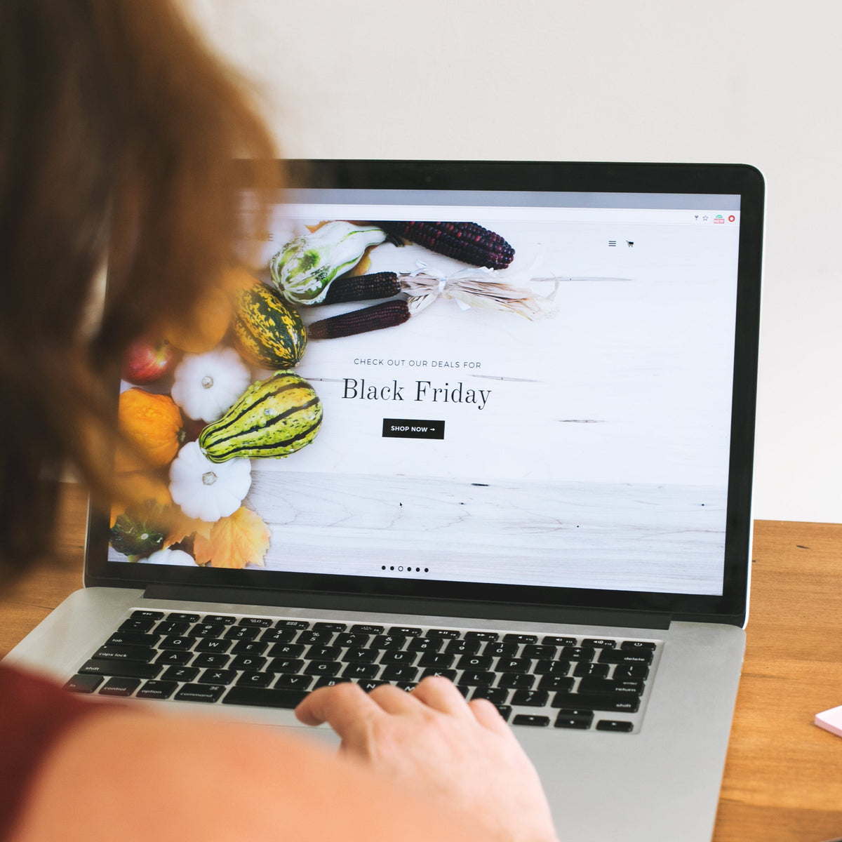 A person viewed from behind sits at a wooden desk using a laptop computer displaying a Black Friday webpage. A white globe and a small pink sticky note sit on the desk next to the laptop against a white wall.